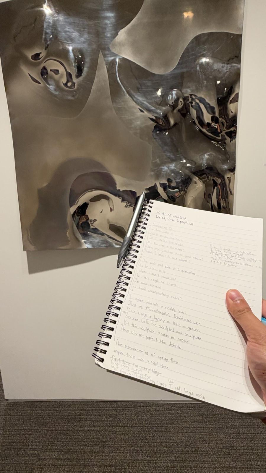 A polished, warped metal wall sculpture at the Ackland Art Museum, with Justin's notebook held up in front of it showing the handwritten first draft of this poem.